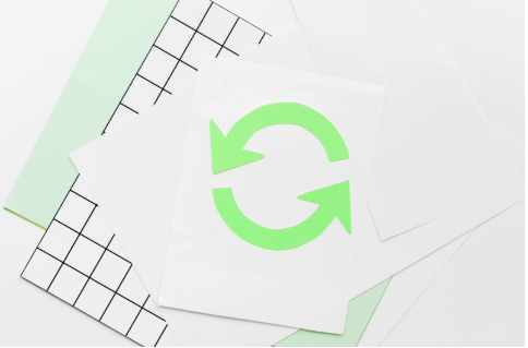 Folha de papel com símbolo verde de reciclagem em setas circulares, sobreposta a outras folhas e fundo geométrico