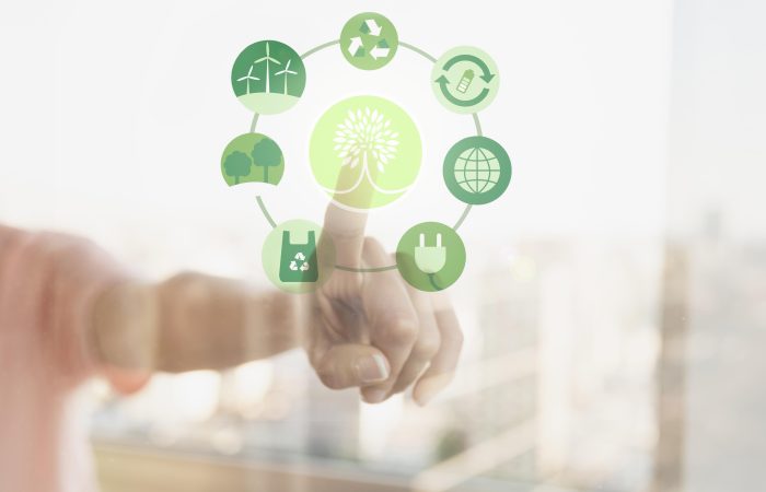 Pessoa tocando em uma tela transparente com ícones verdes relacionados à sustentabilidade, incluindo energia renovável, reciclagem, reutilização, preservação de árvores, sacola reciclável, conexão global e energia limpa, em torno de um símbolo central de árvore estilizada