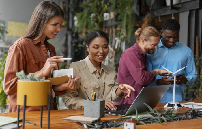 Grupo de jovens profissionais em ambiente colaborativo e sustentável, reunidos em torno de um notebook e um modelo de turbina eólica, discutindo ideias de tecnologia e sustentabilidade