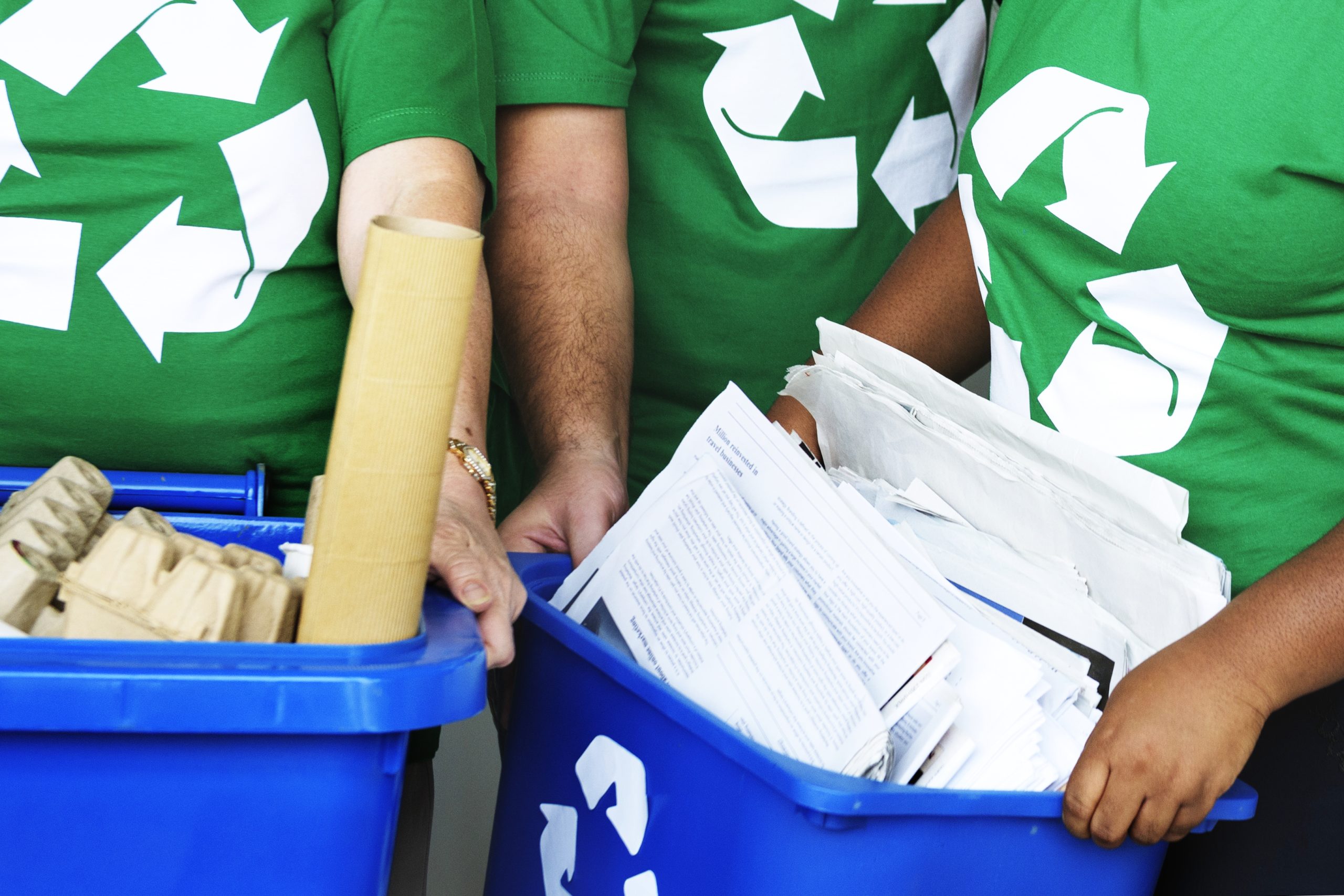 Pessoas com camisetas verdes de reciclagem segurando caixas azuis cheias de papéis e materiais recicláveis, simbolizando a gestão de resíduos e a coleta seletiva.