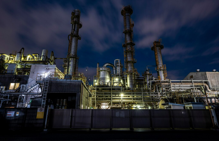 Vista noturna de uma planta industrial com grandes torres de destilação e tubulações iluminadas, representando uma estrutura geradora de resíduos Classe I e Classe II que exige gestão ambiental rigorosa.