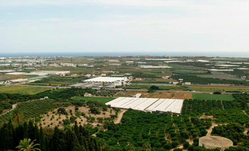 Fotografia aérea ampla de uma vasta região que combina áreas verdes e estruturas de produção, representando um parque de ecologia industrial integrado à paisagem natural. Em primeiro plano, destacam-se plantações, pomares e árvores nativas circundando grandes galpões operacionais horizontais de cor branca com coberturas industriais. Ao longo da extensão, veem-se outras instalações fabris, estufas e vias de circulação integradas, terminando na linha do horizonte que faz limite com uma cidade costeira e o mar azul sob um céu claro. A imagem ilustra o conceito de ecoparque e simbiose industrial em harmonia com o meio ambiente.