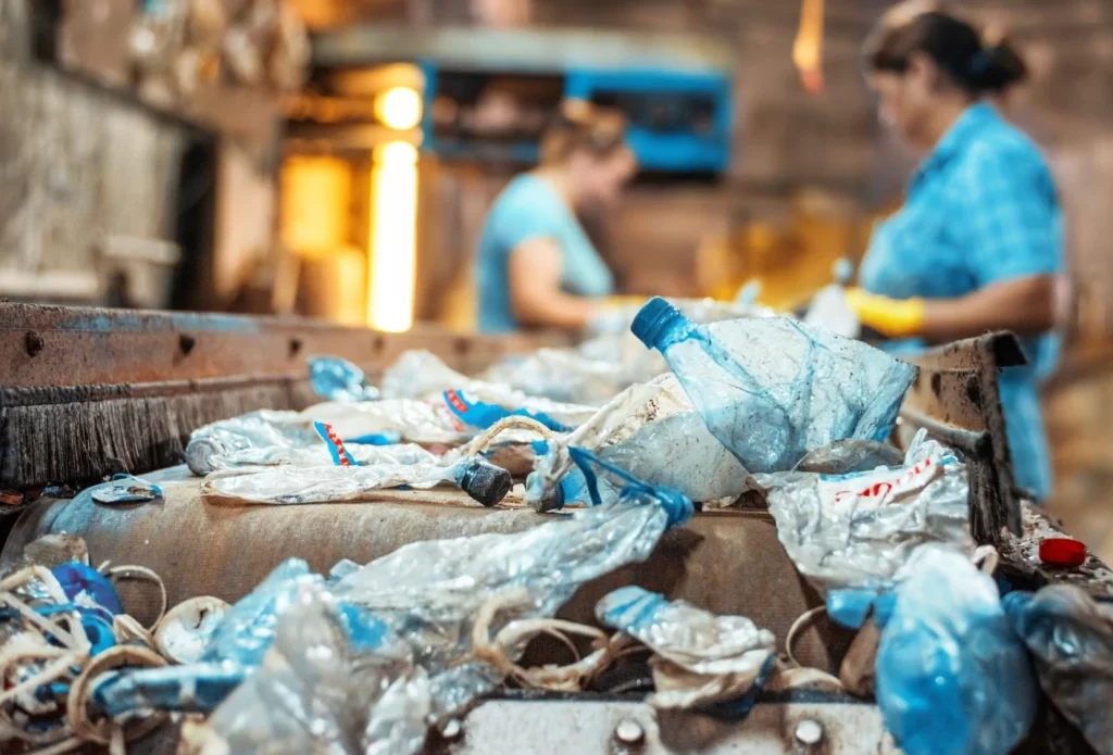 Uma correia transportadora em uma fábrica de reciclagem está repleta de garrafas de plástico amassadas e outros resíduos, com foco em uma garrafa de plástico azul. Ao fundo, fora de foco, duas trabalhadoras operam a linha de triagem. A imagem ilustra o crescimento do setor de gestão de resíduos e a importância da economia circular.